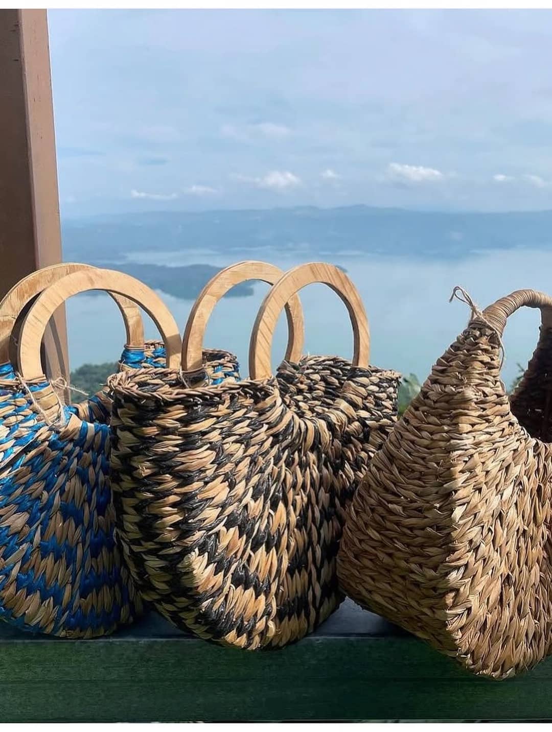 Tres carteras de fibra natural con asas de madera frente al lago.